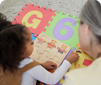 A young child working on a wooden puzzle board with an adult on a colorful foam play mat with letters and numbers
