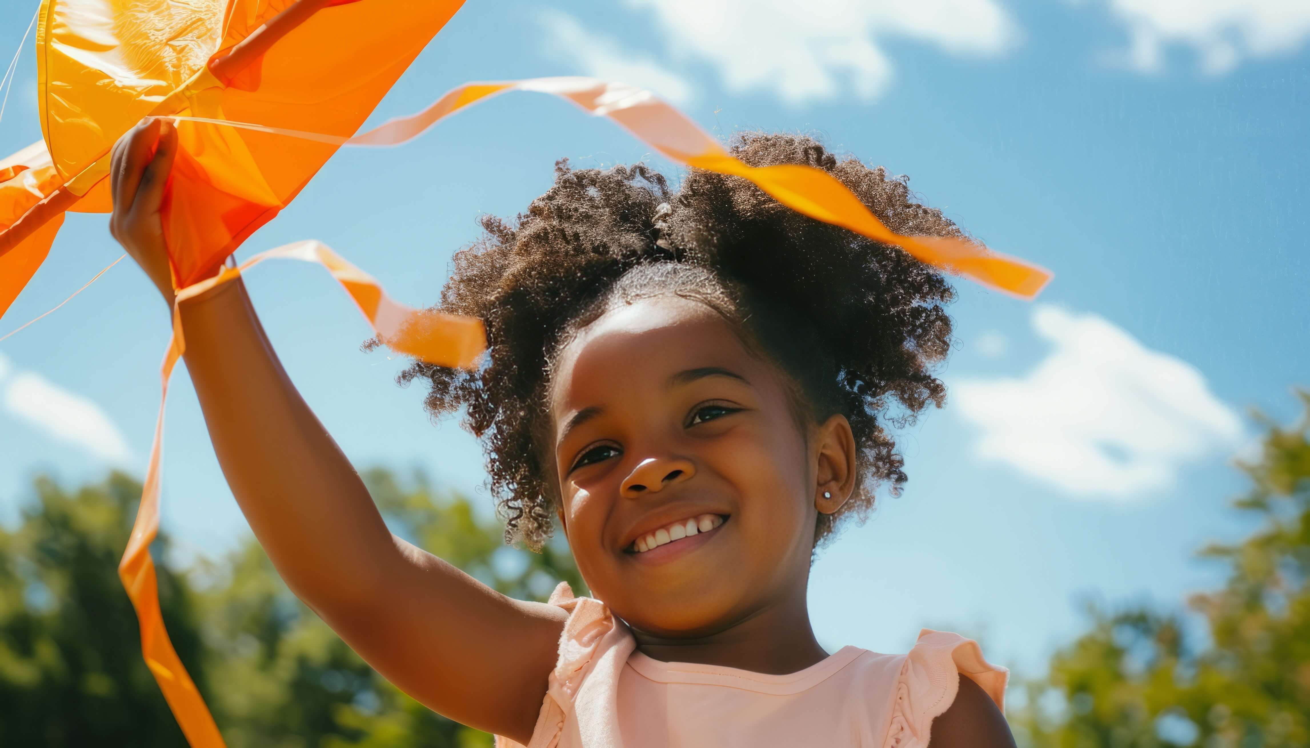 Girl smiling while flying an orange kite on a sunny summer day