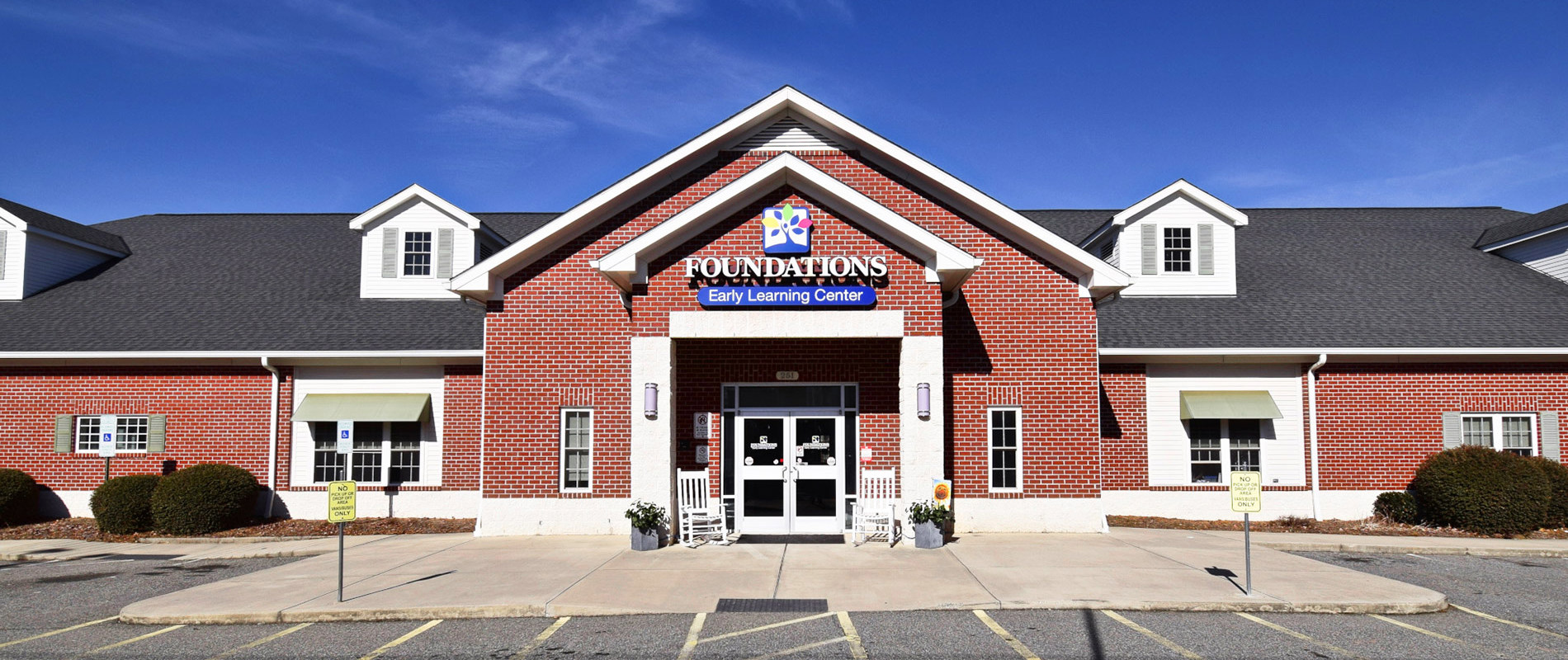 Exterior of Foundations Early Learning Center building with red brick facade, white trim, gabled entrance with school signage, and two white rocking chairs flanking the front door