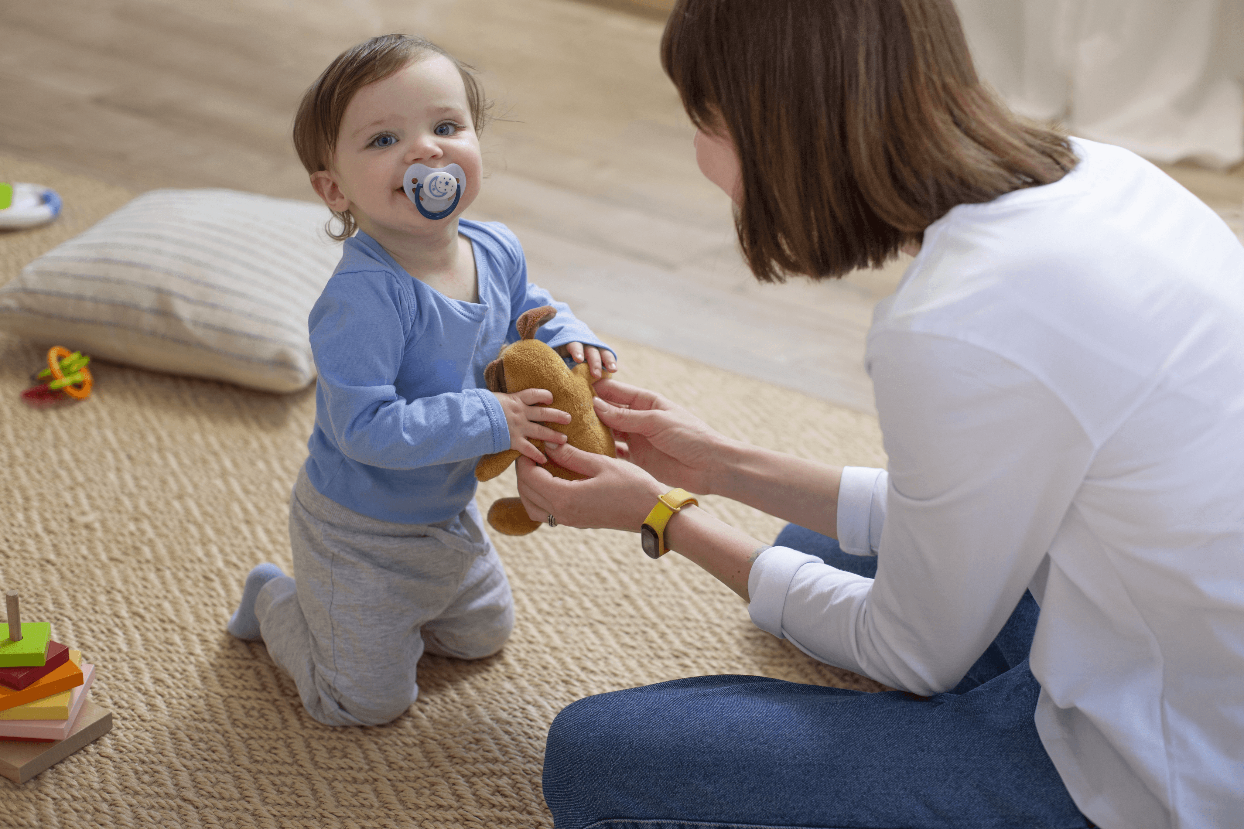 A toddler with a pacifier in a blue outfit holds a stuffed bear while interacting with a caregiver kneeling on the floor