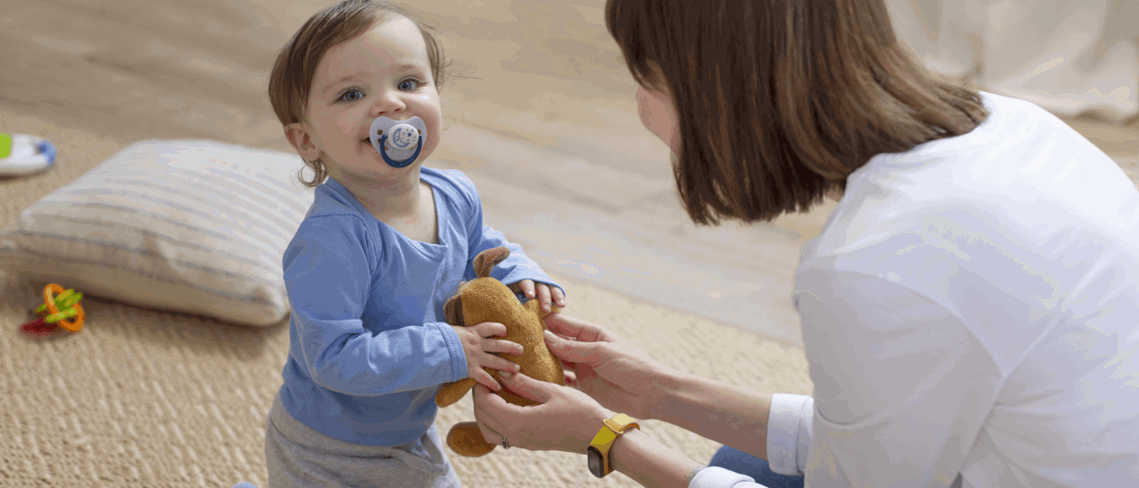 A toddler with a pacifier in a blue outfit holds a stuffed bear while interacting with a caregiver kneeling on the floor