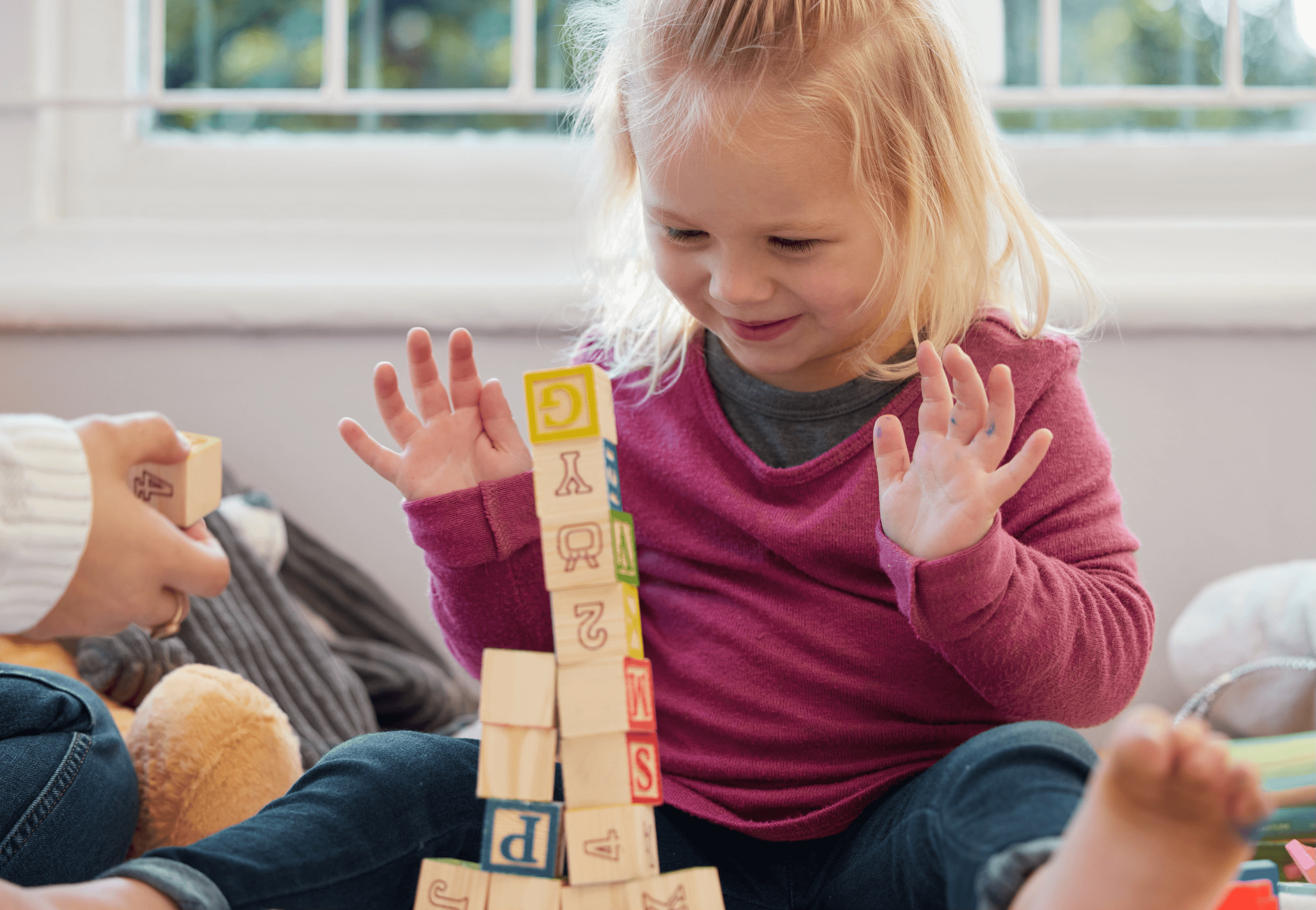 A young toddler girl with blonde hair excitedly watching a tall stack of wooden alphabet blocks, hands raised in delight, sitting on the floor at play