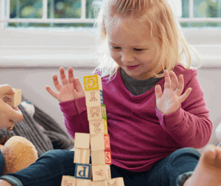 A young toddler girl with blonde hair excitedly watching a tall stack of wooden alphabet blocks, hands raised in delight, sitting on the floor at play