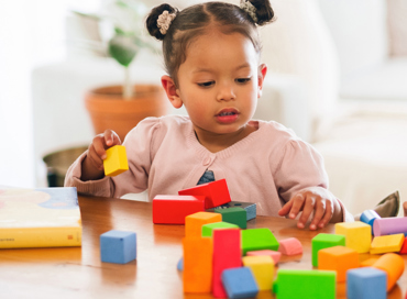 A young toddler girl with hair buns playing with colorful wooden building blocks at a table, representing the Step-Up Early Preschool program