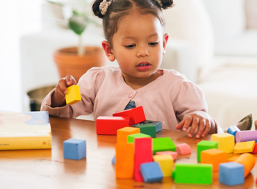 A young toddler girl with hair buns playing with colorful wooden building blocks at a table, representing the Step-Up Early Preschool program