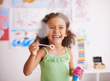 A happy girl with curly hair laughing while blowing bubbles in a bright classroom, representing the before and after school program