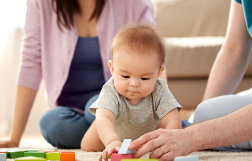 A baby sitting on the floor playing with colorful building blocks, with two adults seated nearby in a home setting