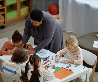 A teacher in a grey dress leans over a table assisting several young children with a coloring and art activity in a bright classroom