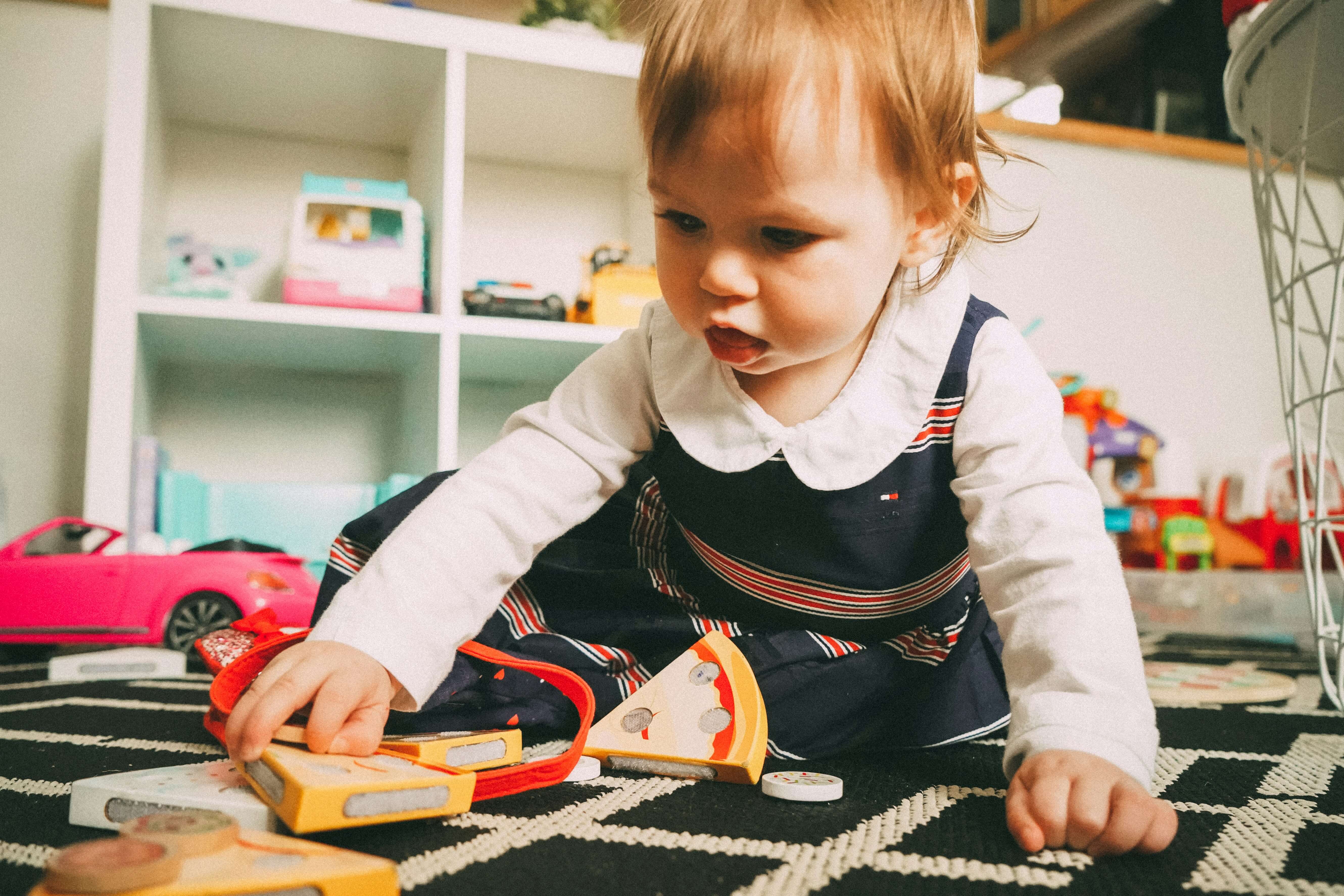 A baby girl in a navy striped dress exploring wooden toys on the floor of a playroom with a toy shelf in the background