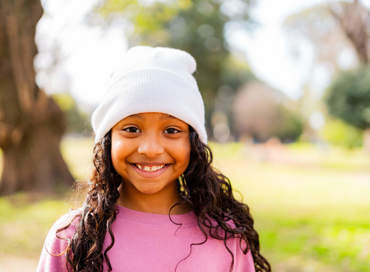 A smiling girl with curly hair wearing a white beanie and pink sweater outdoors in a park, representing the school break care program