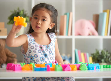 A young girl in a floral dress holding up a yellow building block while playing with colorful toys at a table, representing the preschool program