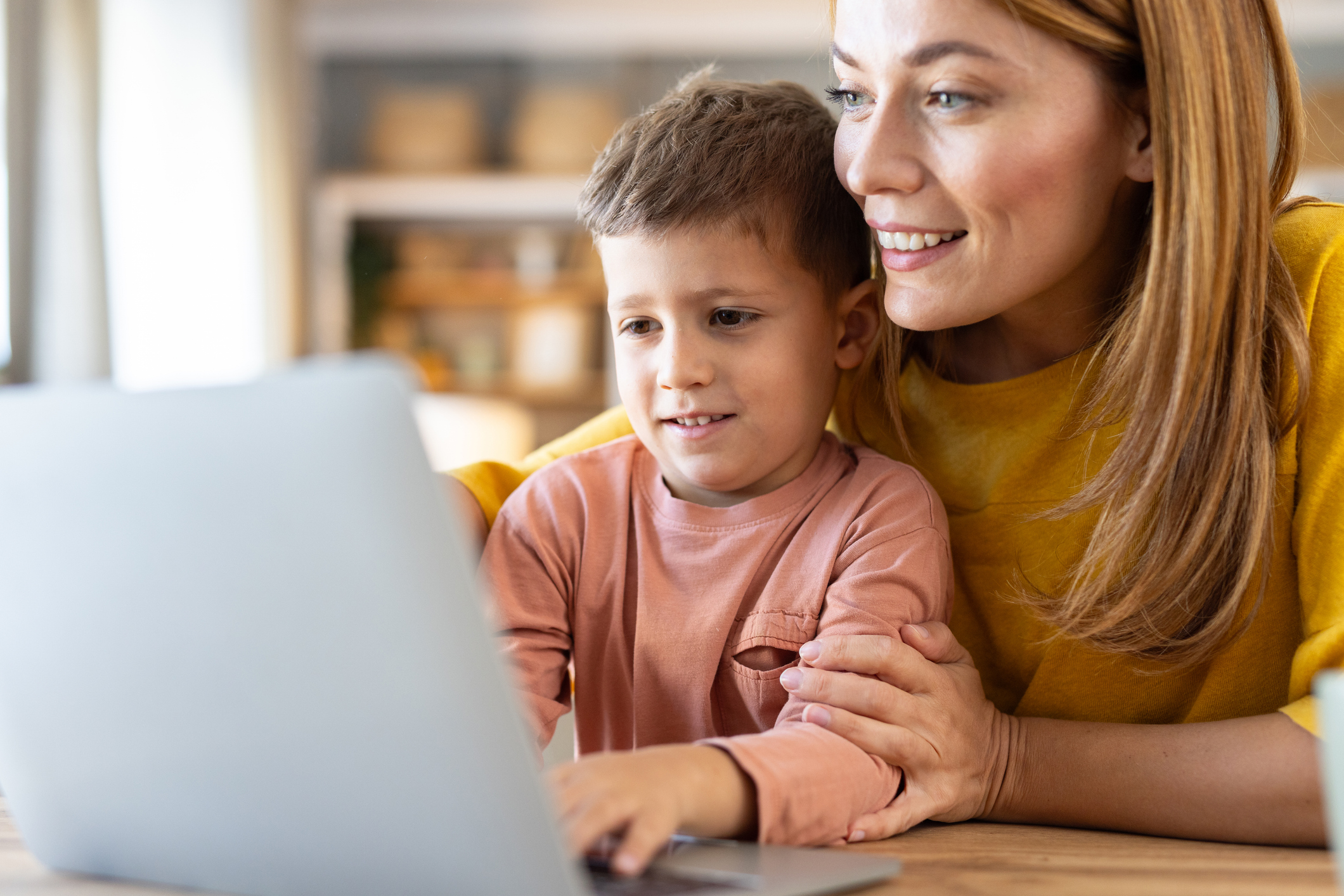 A smiling woman in a yellow sweater and a young boy looking at a laptop screen together at a table