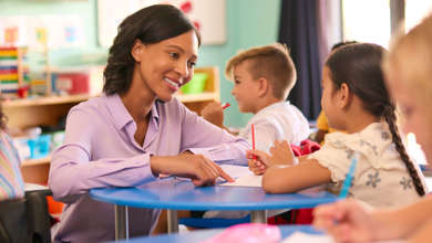 A smiling teacher in a purple shirt leaning toward children seated at a round table in a bright early learning classroom